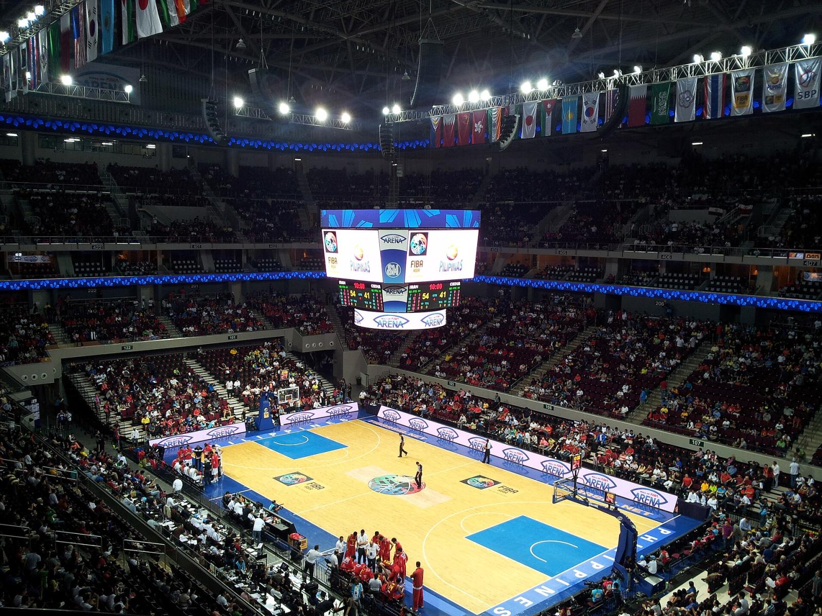 A packed Mall of Asia Arena during an international basketball tournament, with competing nations' flags hung from the roof
